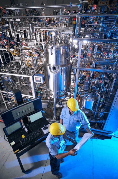 Workers in helmets inspect documents in an industrial facility with complex machinery