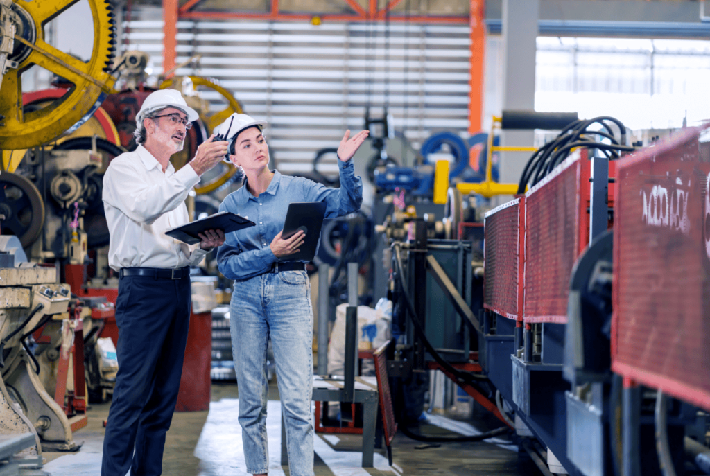 Two people in a factory discussing machinery, one holding a clipboard and the other a tablet